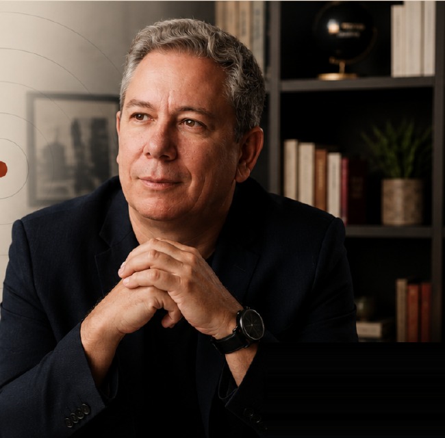 Marcelo Dias sentado, com as mãos unidas, em frente a uma estante de livros